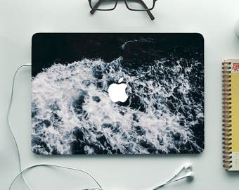 May include: A black laptop with a white wave design on the lid. The laptop is on a white surface with a pair of glasses, a notebook, a pencil, and a cup of coffee.