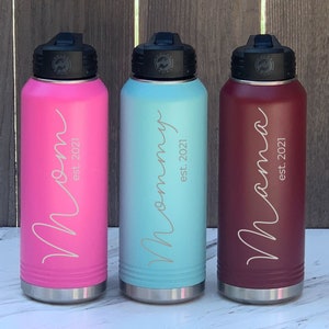 May include: Three stainless steel water bottles with different colored exteriors: pink, light blue, and burgundy. Each bottle has a black lid and a silver base. The bottles are engraved with the words "Mom est. 2021", "Mommy est. 2021", and "Mama est. 2021".