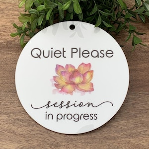 Puede incluir: Un letrero redondo blanco con el texto "Quiet Please" y una ilustración de una flor de loto rosa y amarilla. El texto "session in progress" está escrito en cursiva debajo de la flor.