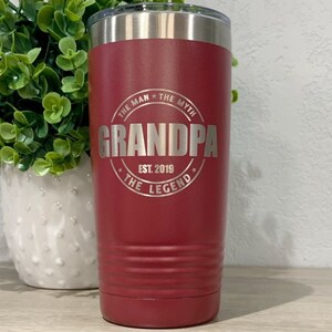 May include: A red stainless steel tumbler with a silver inscription that reads "The Man * The Myth * Grandpa * Est. 2019 * The Legend".