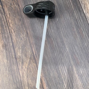 May include: A black plastic bottle lid with a white straw extending from the center. The lid has a gray handle and a textured design.