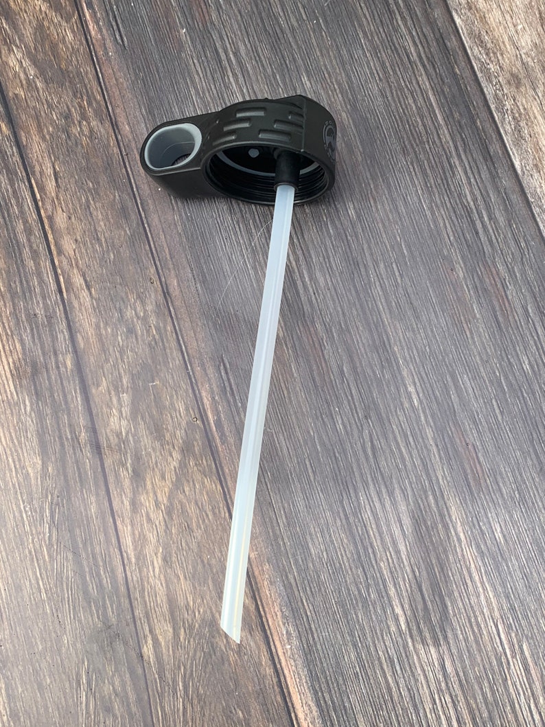 May include: A black plastic bottle lid with a white straw extending from the center. The lid has a gray handle and a textured design.