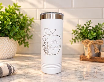 May include: A white cylindrical stainless steel tumbler with a metallic silver lid. The front features a line art graphic of an apple containing a sprig of lavender and the text "Mrs. Raymond" in a cursive script font.