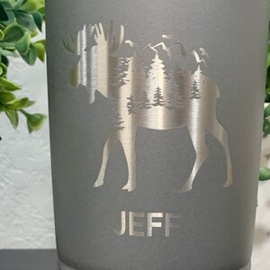 May include: Gray stainless steel tumbler with a silver moose silhouette design. The moose is surrounded by trees and mountains. The name "JEFF" is engraved below the moose.