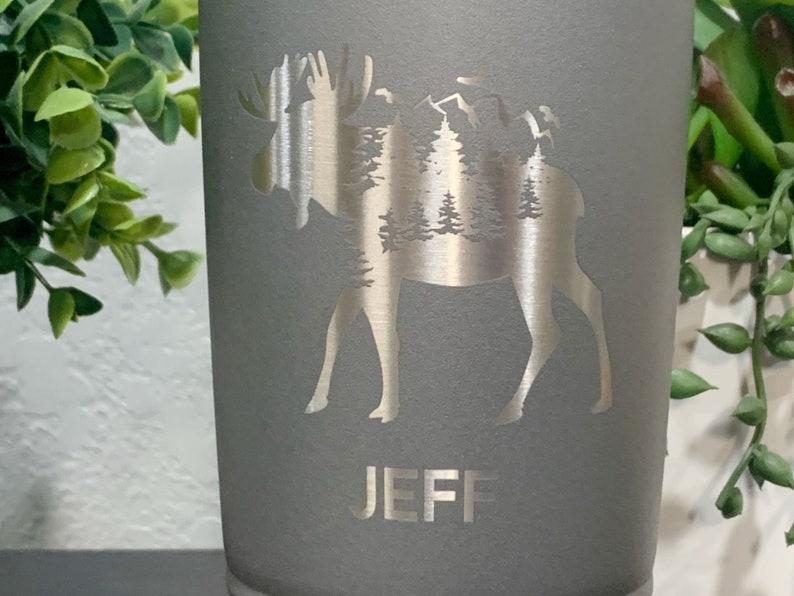 May include: Gray stainless steel tumbler with a silver moose silhouette design. The moose is surrounded by trees and mountains. The name "JEFF" is engraved below the moose.