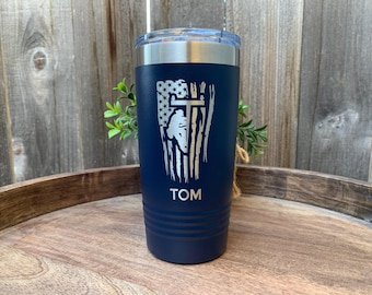 May include: A navy blue stainless steel tumbler with a white and silver design of an American flag with a cross. The tumbler has the name "TOM" engraved on the bottom.