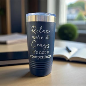 May include: A navy blue insulated tumbler with a silver rim and lid. The tumbler has the words "Relax we're all Crazy it's not a competition" in a white script. The tumbler is on a wooden surface.