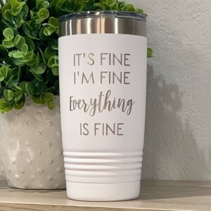 K&ouml;nnte beinhalten: Wei&szlig;er isolierter Becher mit silbernem Deckel. Der Becher hat den eingravierten Schriftzug "IT'S FINE I'M FINE Everything IS FINE". Der Becher hat ein geriffeltes Design in der N&auml;he des Bodens.