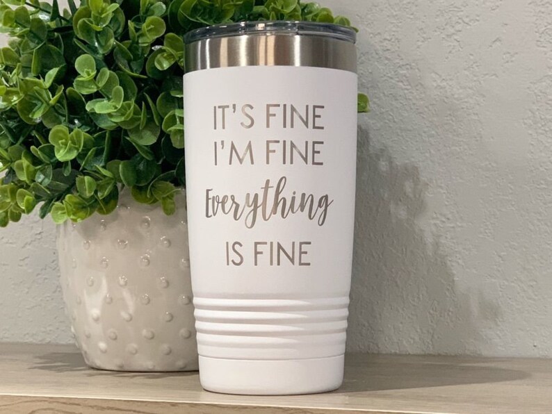 K&ouml;nnte beinhalten: Wei&szlig;er isolierter Becher mit silbernem Deckel. Der Becher hat den eingravierten Schriftzug "IT'S FINE I'M FINE Everything IS FINE". Der Becher hat ein geriffeltes Design in der N&auml;he des Bodens.