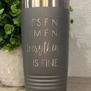 K&ouml;nnte beinhalten: Ein grauer Isolierbecher mit silbernem Rand und Deckel. Der Becher tr&auml;gt den eingravierten Text "IT'S FINE, I'M FINE, Everything IS FINE". Der Becher ist zylindrisch mit horizontalen Rillen.