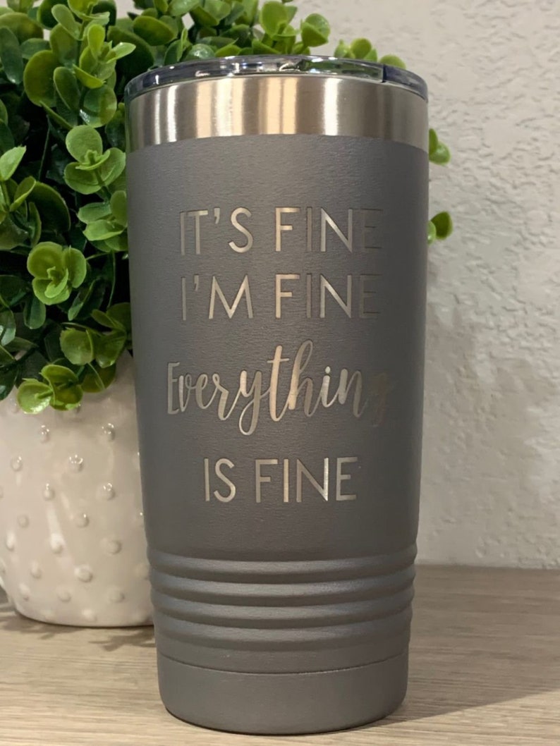 K&ouml;nnte beinhalten: Ein grauer Isolierbecher mit silbernem Rand und Deckel. Der Becher tr&auml;gt den eingravierten Text "IT'S FINE, I'M FINE, Everything IS FINE". Der Becher ist zylindrisch mit horizontalen Rillen.