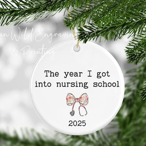 May include: A white ceramic Christmas ornament with the text "The year I got into nursing school" and the year 2025. A bow and stethoscope graphic is below the text. The ornament hangs from a gold string.