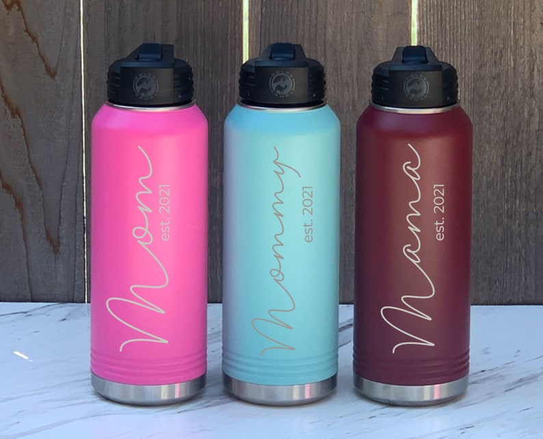 May include: Three stainless steel water bottles with different colored exteriors: pink, light blue, and burgundy. Each bottle has a black lid and a silver base. The bottles are engraved with the words "Mom est. 2021", "Mommy est. 2021", and "Mama est. 2021".