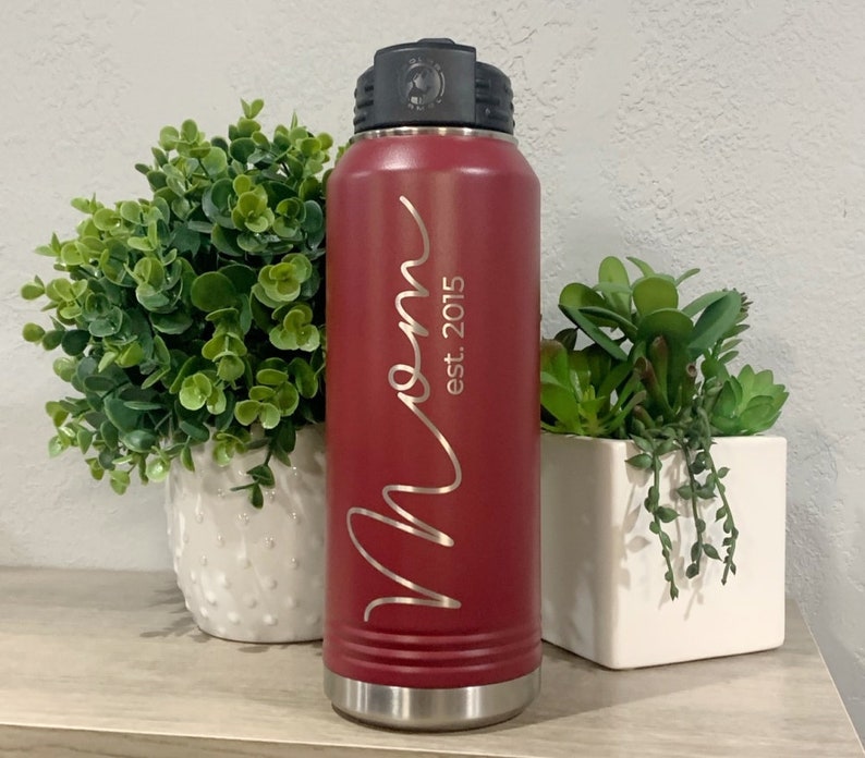 May include: A red stainless steel water bottle with a black lid and a silver base. The bottle has the word "Mom" written in cursive with "est. 2015" underneath.