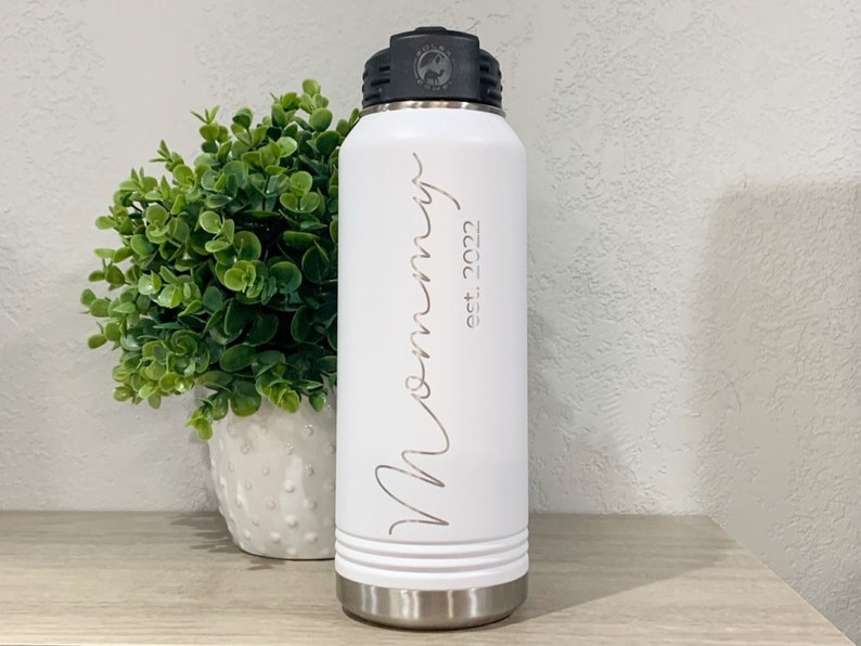 May include: White stainless steel water bottle with a black lid and a white inscription that reads "Mommy est. 2022".