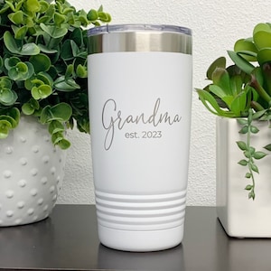 May include: A white insulated tumbler with a silver lid. The tumbler has the word "Grandma" in a script font, with "est. 2023" below it. The tumbler has a ridged design near the bottom.