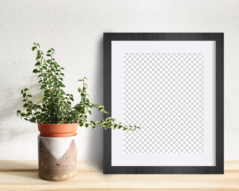May include: A black picture frame with a white mat, sitting on a wooden surface next to a potted plant with green and white leaves. The plant is in a brown and white ceramic pot with a cartoon face.