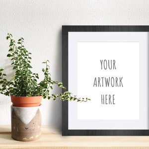 May include: A black picture frame with a white mat and the text "YOUR ARTWORK HERE" on a white background. A potted plant with green and white leaves is in the foreground.