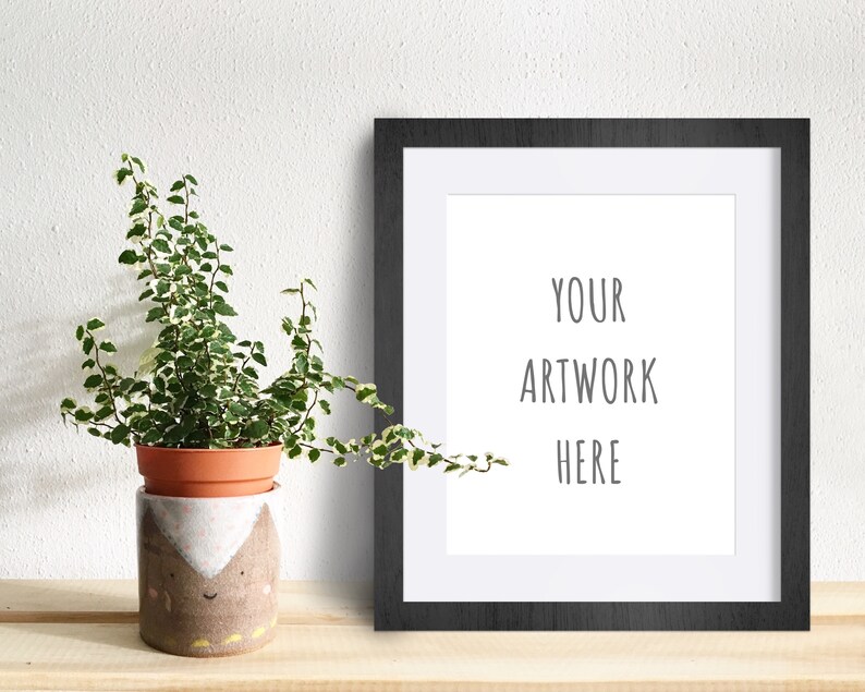May include: A black picture frame with a white mat and the text "YOUR ARTWORK HERE" on a white background. A potted plant with green and white leaves is in the foreground.