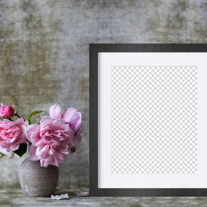 May include: A black picture frame with a white mat sits on a textured gray surface. A vase with pink roses sits in front of the frame.