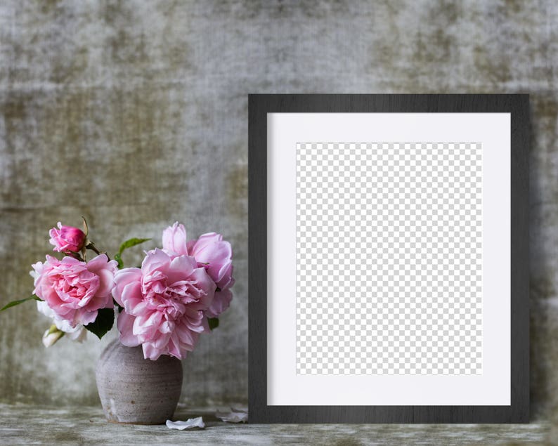 May include: A black picture frame with a white mat sits on a textured gray surface. A vase with pink roses sits in front of the frame.