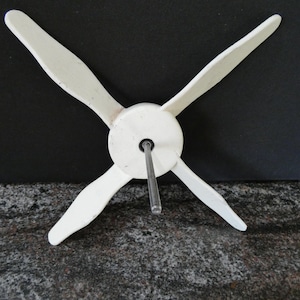 May include: A white, four-bladed propeller with a central hub and a metal rod extending from the centre. The propeller blades are curved and angled, suggesting a wind-powered device. The background is dark.