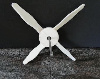 May include: A white, four-bladed propeller with a central hub and a metal rod extending from the center. The propeller blades are curved and angled, suggesting a wind-powered device. The background is dark.