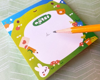 May include: A green frog-shaped notepad with a white writing area and colorful cartoon illustrations around the border. The notepad says "notes" in a circle at the top. A pencil is resting on the notepad with the word "hi" written on the page.