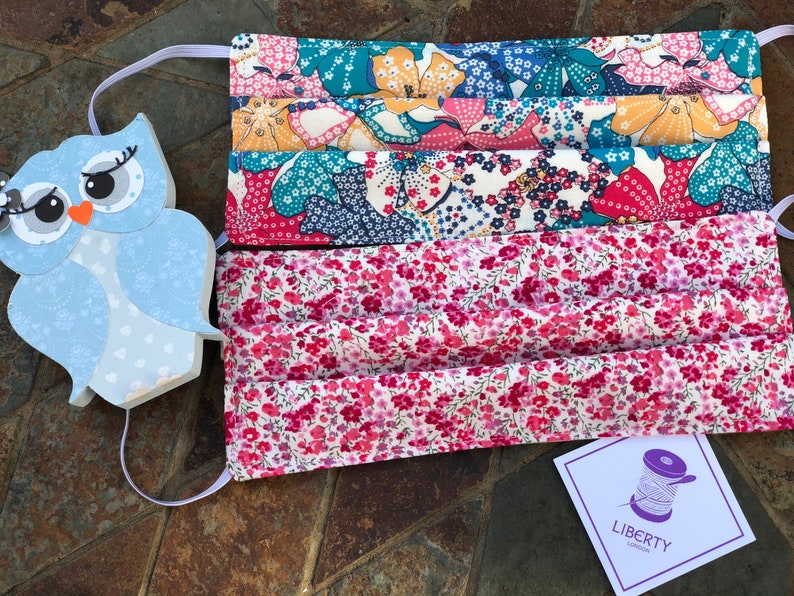 May include: A set of three face masks made from Liberty of London fabric. The masks are pleated and have a floral print in shades of pink, blue and yellow. The masks are made with a white background and have a Liberty of London label attached.