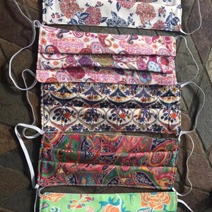 May include: Five different patterned face masks with white elastic ear loops. The masks are made of fabric and have a pleated design. The patterns include floral, paisley, and geometric designs.