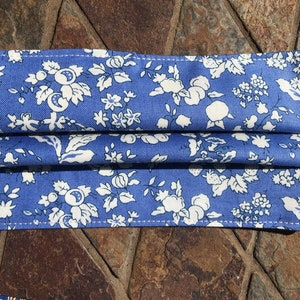 May include: A blue fabric face mask with a white floral pattern. The mask has pleats and elastic ear loops.
