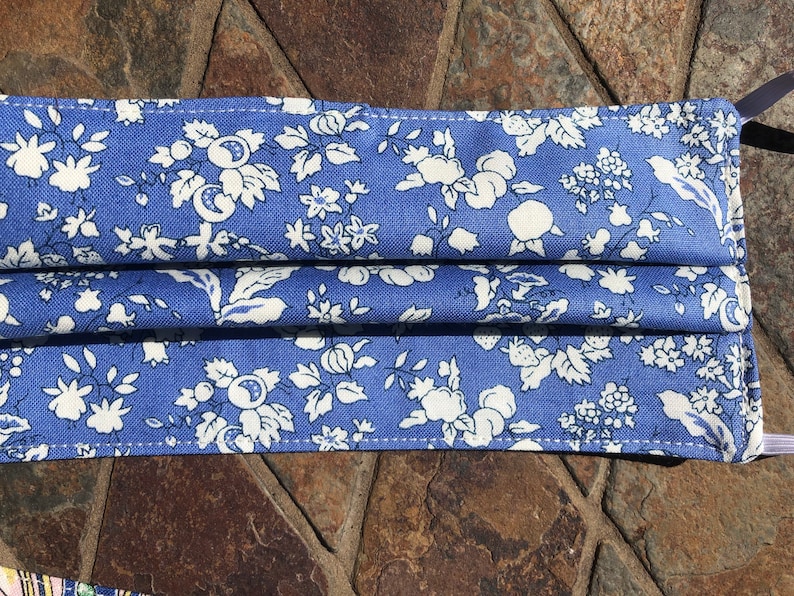 May include: A blue fabric face mask with a white floral pattern. The mask has pleats and elastic ear loops.