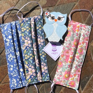 May include: Two face masks with floral patterns and white elastic ear loops. The masks are made with Liberty Fabrics and feature a blue and pink floral pattern and a pink and white floral pattern. The masks are displayed on a stone surface with a small owl cutout and a tag that reads "LIBERTY LONDON".