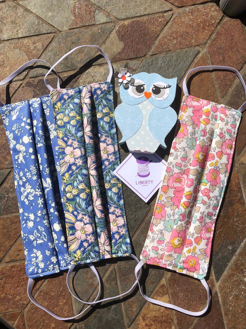 May include: Two face masks with floral patterns and white elastic ear loops. The masks are made with Liberty Fabrics and feature a blue and pink floral pattern and a pink and white floral pattern. The masks are displayed on a stone surface with a small owl cutout and a tag that reads "LIBERTY LONDON".