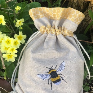 May include: A small, beige drawstring bag with a yellow honeycomb pattern around the top. The bag has a bee embroidered on the front.