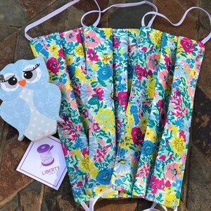May include: Three floral print face masks with white elastic ear loops. The masks are folded and stacked on top of each other. The fabric has a light blue background with pink, yellow, and green flowers. The masks are made by Liberty London.