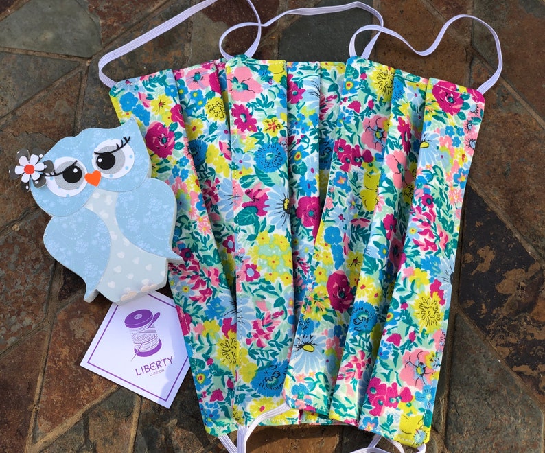 May include: Three floral print face masks with white elastic ear loops. The masks are folded and stacked on top of each other. The fabric has a light blue background with pink, yellow, and green flowers. The masks are made by Liberty London.