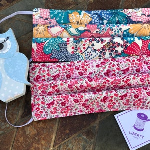 May include: A set of three face masks made from Liberty of London fabric. The masks are pleated and have a floral print in shades of pink, blue and yellow. The masks are made with a white background and have a Liberty of London label attached.