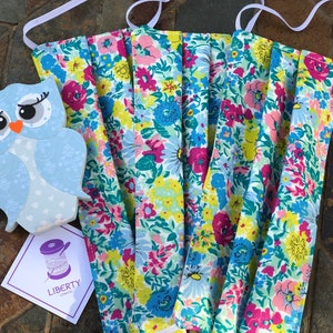 May include: Five face masks with a floral pattern in shades of pink, yellow, blue, and green. The masks are made of fabric and have white elastic ear loops. The masks are arranged in a stack, with the top mask folded in half. The masks are sitting on a brown tiled surface. A small blue owl figurine and a white card with the text "LIBERTY LONDON" are also visible in the image.