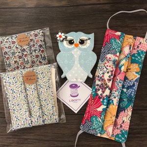 May include: Three pieces of fabric with floral patterns, two with a brown background and one with a white background. The fabric is packaged in clear plastic bags with brown paper tags that say "Handmade with Love". A blue and white owl figurine is in the background. A floral fabric face mask is in the foreground.