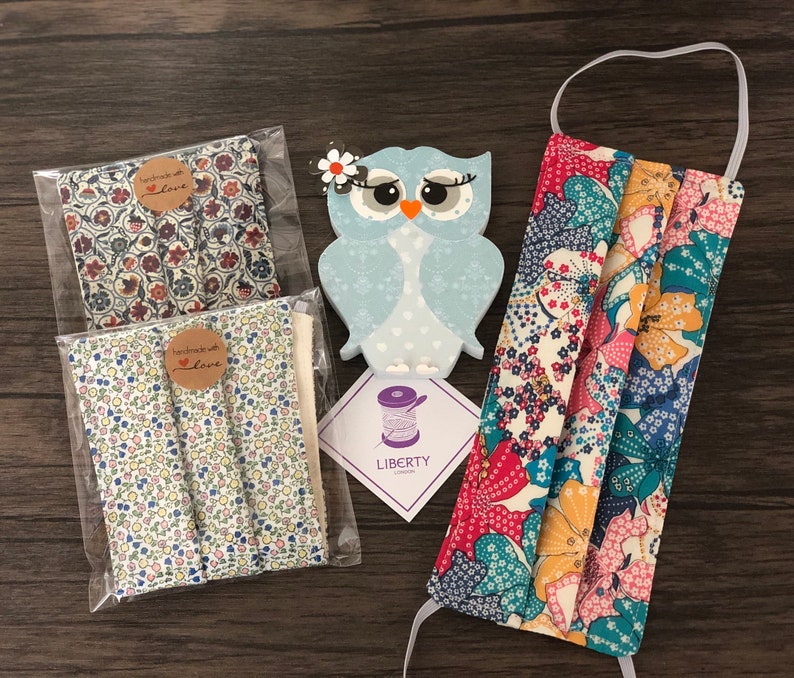 May include: Three pieces of fabric with floral patterns, two with a brown background and one with a white background. The fabric is packaged in clear plastic bags with brown paper tags that say "Handmade with Love". A blue and white owl figurine is in the background. A floral fabric face mask is in the foreground.