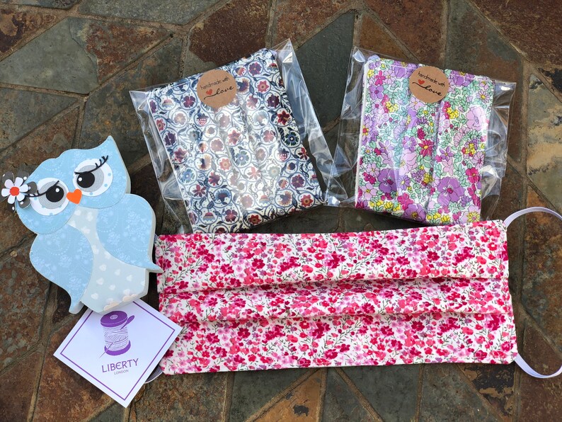 May include: A pink and white floral fabric face mask with a light purple elastic band. The mask is handmade and packaged with a small tag that says "Handmade with Love". Two additional pieces of fabric are also included, one with a blue and white floral pattern and one with a purple and white floral pattern.