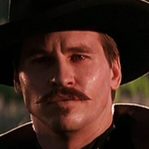 May include: A man wearing a black hat and a black jacket with a white collar. He has a mustache and a serious expression on his face.