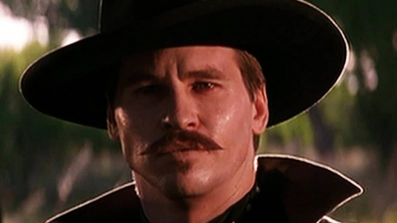 May include: A man wearing a black hat and a black jacket with a white collar. He has a mustache and a serious expression on his face.