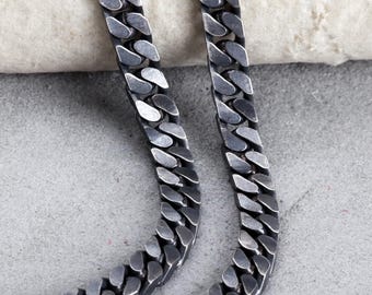 May include: Close-up of two silver-toned curb chain necklaces. The chains are flat and have a series of interlocking links. The necklaces are displayed on a light-colored surface.