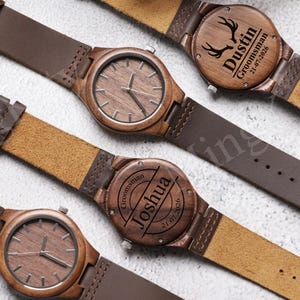 K&ouml;nnte beinhalten: Vier Holzuhren mit braunen Lederarmb&auml;ndern. Die Zifferbl&auml;tter der Uhren sind holzfarben mit einfachen Zeigern. Zwei Uhren haben personalisierte Gravuren auf der R&uuml;ckseite, einschlie&szlig;lich Namen und Daten. Die Uhren liegen auf einer hellen Oberfl&auml;che.