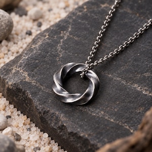 May include: A dark silver-toned metal pendant shaped as a twisted circular ring, suspended from a matching dark silver-toned metal chain. The pendant features a smooth, polished finish with darkened recessed areas that highlight the spiral design.