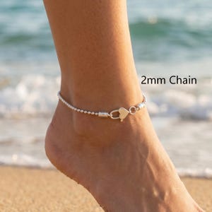 May include: A silver anklet featuring a 2mm chain with a heart-shaped clasp. The jewelry piece is displayed with the text 2mm Chain visible nearby. The design consists of small spherical beads connected in a continuous line around the ankle.