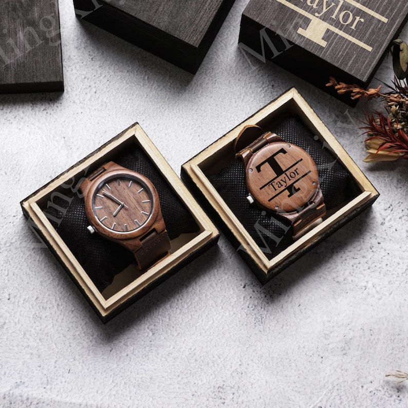 K&ouml;nnte beinhalten: Zwei Holzuhren mit braunen Lederarmb&auml;ndern, pr&auml;sentiert in einzelnen Holzboxen. Eine Uhr hat ein schlichtes Zifferblatt, die andere zeigt den Buchstaben "T" und den Namen "Taylor" eingraviert. Die Boxen sind mit schwarzem Stoff ausgekleidet.