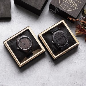 May include: Two wooden watches in open boxes. One watch has a dark wood face and the other has the name "Joshua" engraved on the face. The boxes are dark wood with a light wood frame.